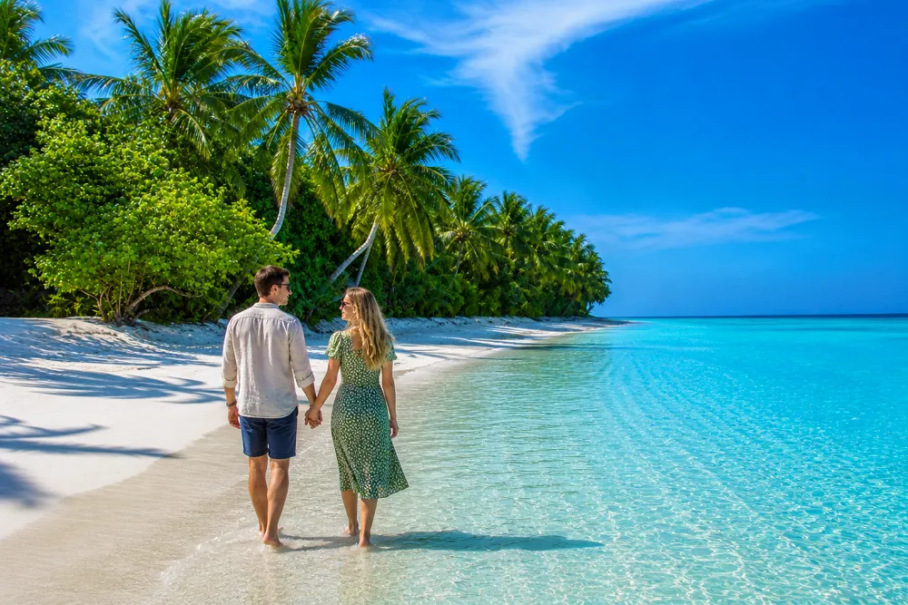 A couple walking hand in hand along a tropical white-sand beach with crystal-clear turquoise water, palm trees, and a bright blue sky.