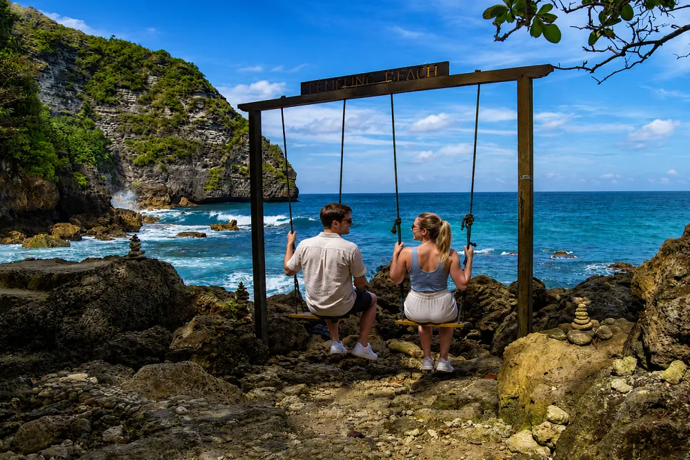 A couple sitting on wooden swings overlooking a tropical rocky beach with turquoise ocean water, cliffs, and a bright blue sky.