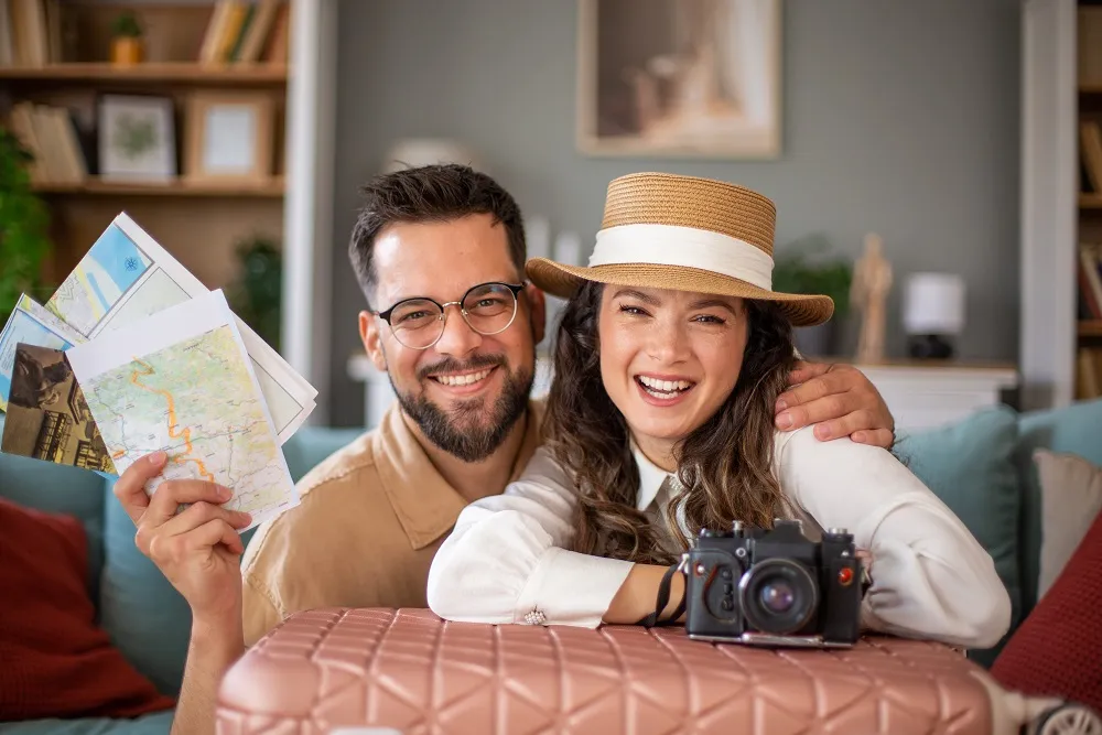 A smiling couple sitting indoors with travel maps, a camera, and a suitcase while planning their next holiday together.
