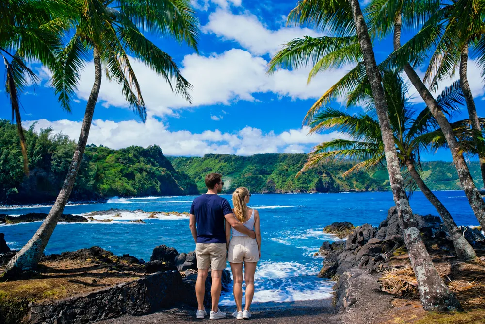 A couple standing together on a tropical coastline surrounded by palm trees, turquoise ocean water, rocky shores, and lush green cliffs under a bright blue sky.