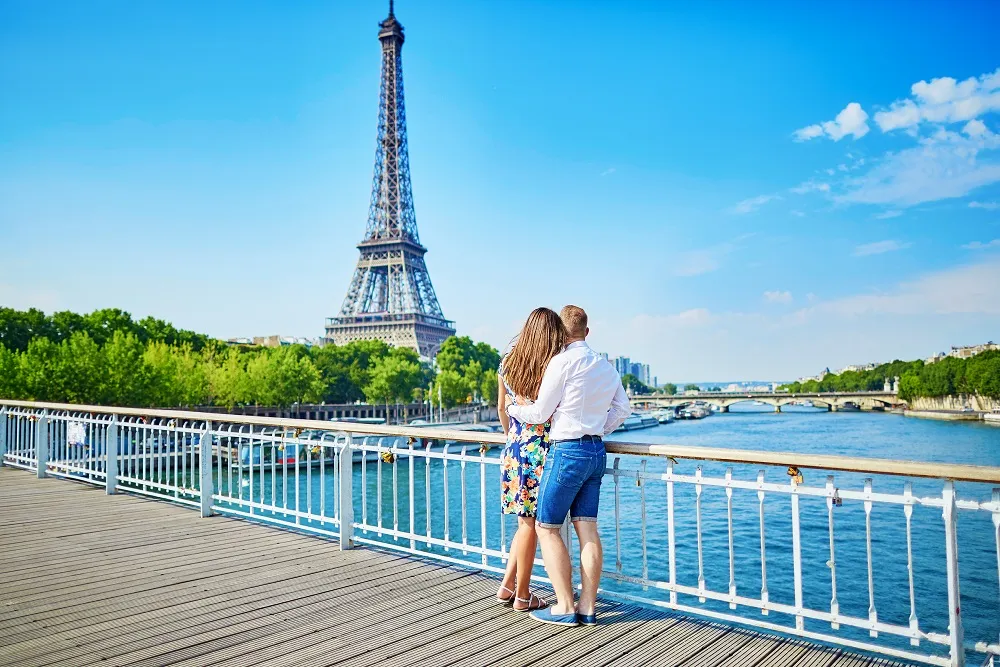 A couple embracing on a riverside walkway in Paris with the Eiffel Tower in the background on a bright sunny day beside the River Seine.