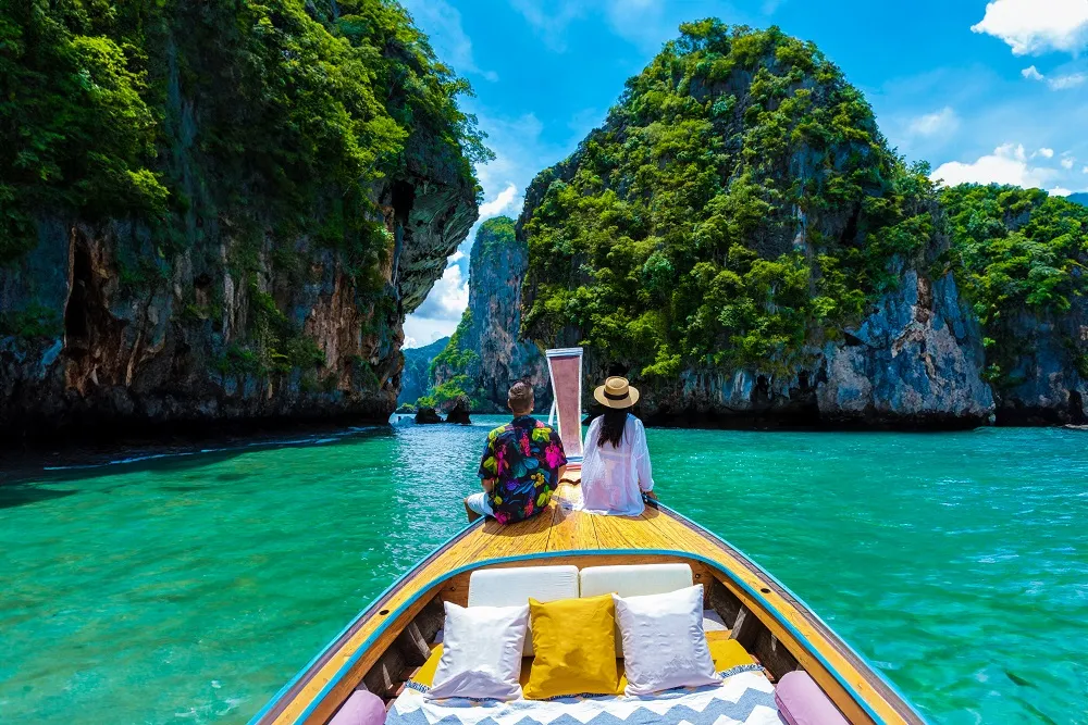 A couple sitting on the front of a wooden boat cruising through turquoise tropical waters surrounded by lush green limestone cliffs under a bright blue sky.