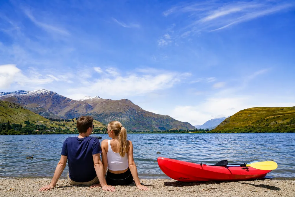A couple sitting beside a lake in Queenstown, New Zealand, with a red kayak, clear blue water, green hills, and snow-capped mountains in the background.